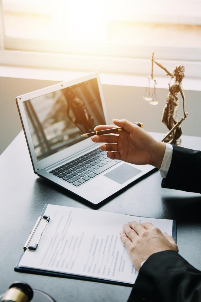 Eine Person sitzt an einem Schreibtisch, auf dem ein Laptop, eine Justitia-Figur und Dokumente auf einem Klemmbrett liegen. Die Person h&auml;lt einen Stift in der Hand und zeigt auf das Klemmbrett. Im Hintergrund ist ein Fenster mit einfallendem Sonnenlicht zu sehen.