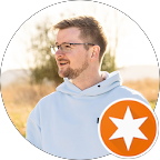 Ein Mann mit Brille und Bart trägt einen hellblauen Kapuzenpullover und blickt nach links. Unten rechts im Bild befindet sich ein Symbol mit einem orangefarbenen Kreis und einem weißen Stern. Der Hintergrund zeigt unscharfe Bäume und einen hellen, sonnigen Himmel.